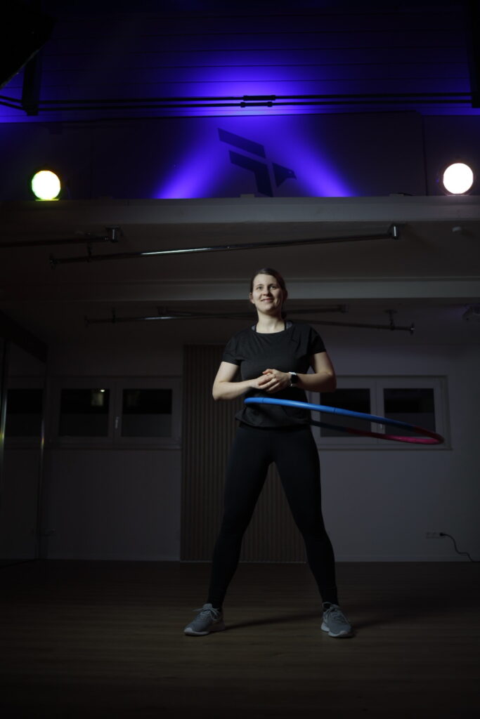 Simone führt einen Hula-Hoop-Kurs im Fitnessstudio und motiviert die Teilnehmer zu spielerischer Bewegung.