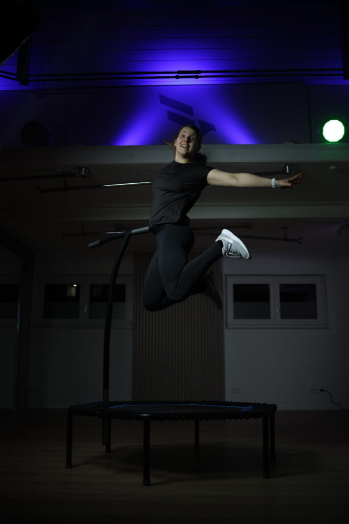 Simone springt mit Energie auf einem Fitness-Trampolin im Studio, umgeben von dramatischer Beleuchtung.