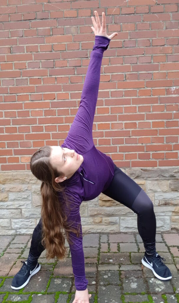 Simone führt eine Dehnübung im Ausfallschritt an einer Backsteinmauer im Freien aus, mit einer Hand am Boden und der anderen in die Luft gestreckt.